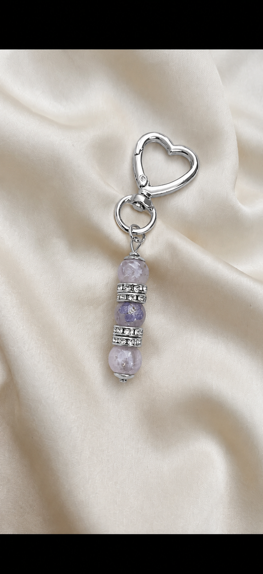 A silver-toned heart-shaped clasp keychain or bag charm with three lavender-purple gemstone beads and silver spacer rings, displayed on a draped cream satin fabric background.
