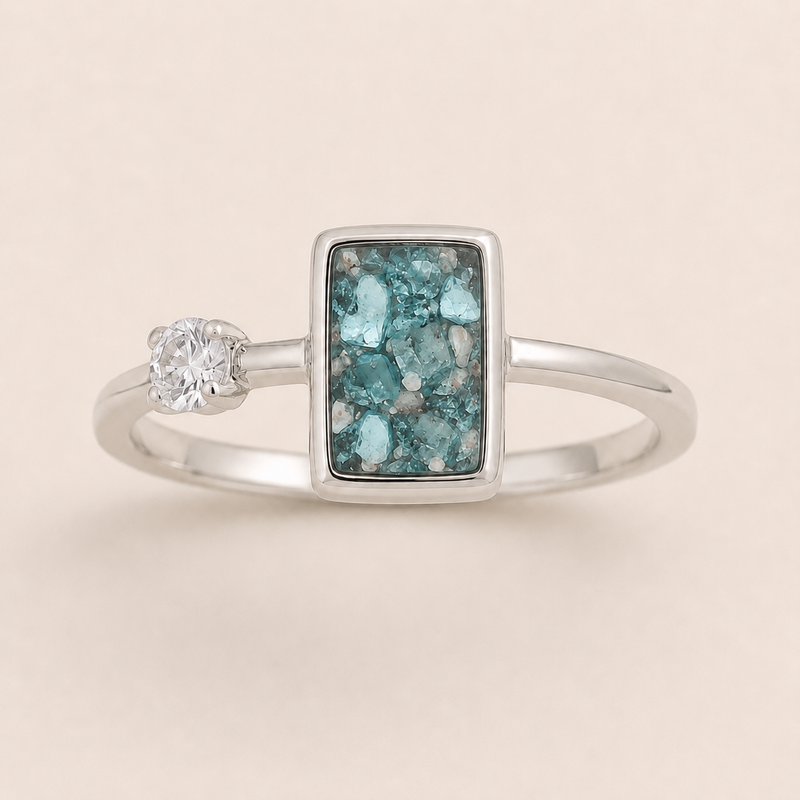 A silver ring featuring a rectangular turquoise druzy stone centerpiece and a small round clear gemstone on the band, set against a solid light beige background.