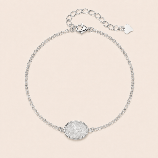 A delicate silver chain bracelet with a textured oval centerpiece and an adjustable lobster clasp with an extension chain and small heart charm, displayed on a light beige background.