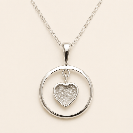 A silver necklace featuring a circular open pendant with a small, sparkling heart-shaped charm in the center, suspended from a delicate silver chain against a light beige background.