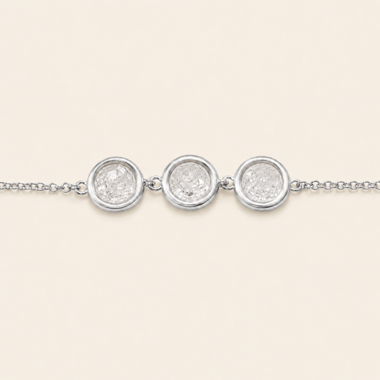 A delicate silver chain bracelet featuring three round bezel-set clear stones, presented on a solid light cream background.