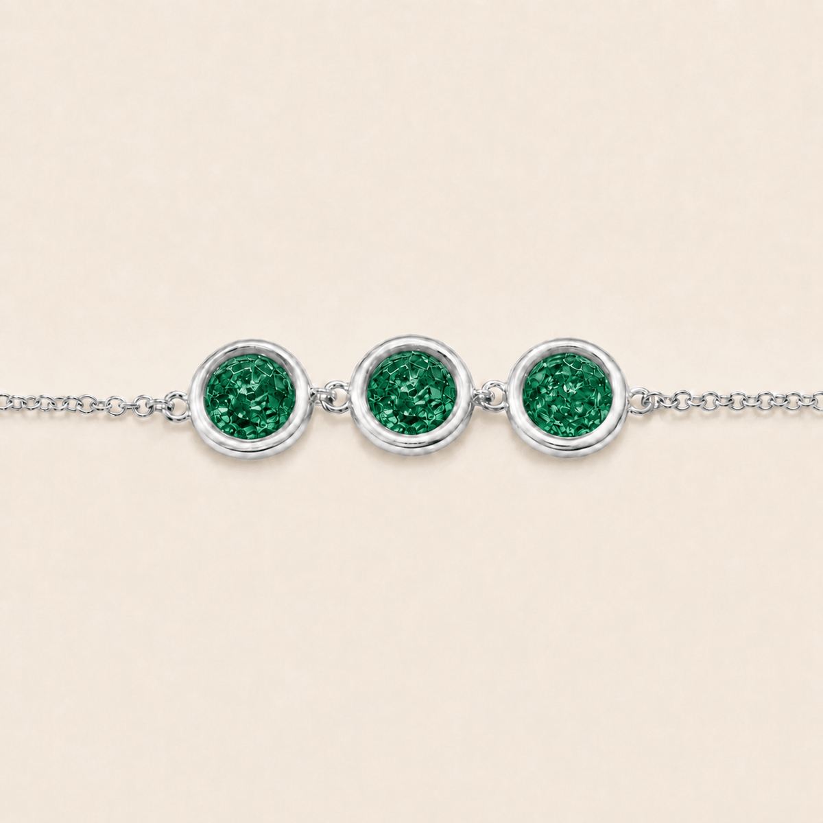 A silver chain bracelet featuring three round bezel-set emerald-cut green gemstones, displayed on a light beige background.