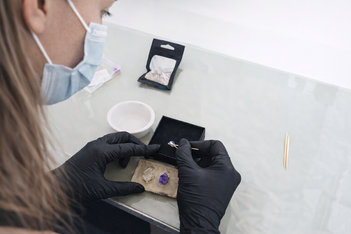 Person wearing gloves and a mask examining jewelry on a table.